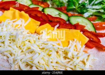 Colorful strips of fresh vegetables including red peppers, cucumbers, and lettuce are lined up next to shredded cheese on a tortilla, ready for wrappi Stock Photo