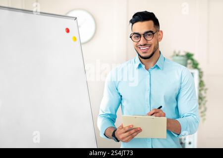 Positive guy English teacher explaining foreign language rules near blackboard, taking notes in clipboard at office Stock Photo