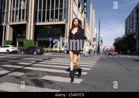 Stylish Young Fashion Woman With Long Hair Walking Across Pedestrian Crossing In City, Wearing Black Mini Dress And Boots, Trendy Urban Street Style M Stock Photo