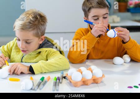 Two brothers are decorating white eggs for easter using colorful markers, sitting at a white table Stock Photo