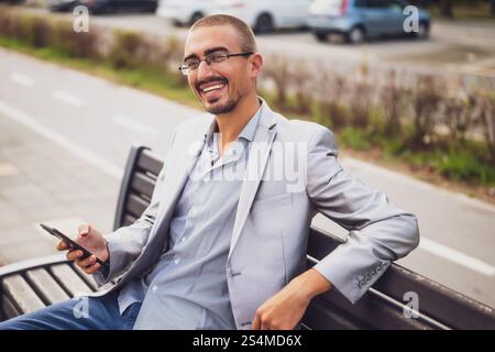 Portrait of cheerful businessman who is messaging on phone. Young businessman sitting on bench in the street and using smartphone. Stock Photo