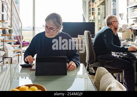 A senior couple sits in a bright home office; one using a tablet, the other focusing on a computer Books and natural light create a cozy, productive a Stock Photo