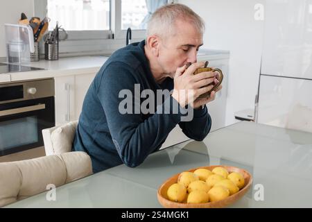 A senior man enjoys a warm cup of coffee at a glass table in a modern kitchen A bowl of lemons decorates the table, adding a pop of color to the seren Stock Photo