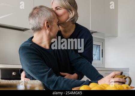 A senior couple enjoys a tender moment in their kitchen One person kisses the other on the head, creating a warm and loving atmosphere A bowl of orang Stock Photo