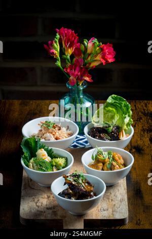 A selection of vibrant Asian cuisine bowls arranged on a wooden board The dishes feature vegetables, meats, and garnishes A vase of bright red flowers Stock Photo