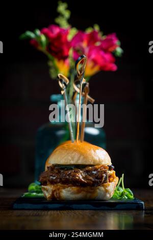 A gourmet pulled pork slider presented elegantly with colorful flowers in the background The food is garnished on a slate, creating an exquisite prese Stock Photo