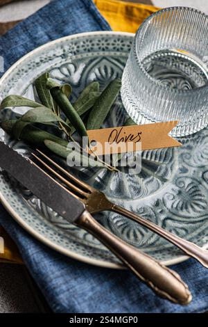 Sophisticated table setting featuring olive leaves on a ceramic plate with a tag, accompanied by a glass and fine dining utensils. Stock Photo
