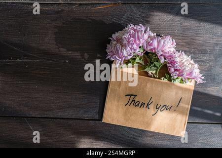 A brown envelope with Thank you written on it holds pink chrysanthemums The flowers rest gracefully on a dark wooden surface, complimenting the fall a Stock Photo