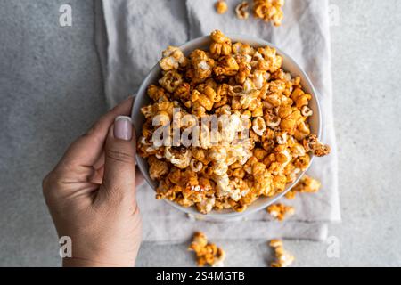 Cropped unrecognizable person holding a bowl of sweet caramel popcorn in a top view, displaying the texture and color of the snack against a muted gra Stock Photo