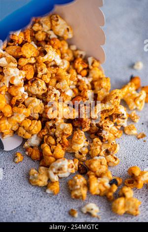 Caramel-coated popcorn spills from a blue container onto a textured gray surface, showcasing the crunchy treat's varied golden hues and textured piece Stock Photo