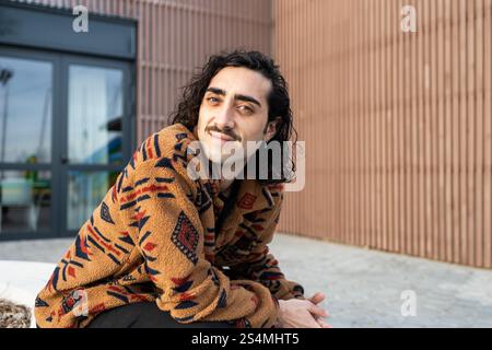 A man with long hair and a mustache, wearing a cozy patterned jacket, smiles while sitting outdoors The background shows a modern building facade, add Stock Photo