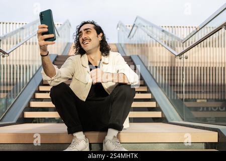 A young man with curly hair and a mustache wearing casual attire sits on outdoor steps, holding a smartphone and taking a selfie The environment is mo Stock Photo