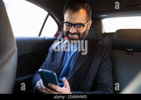A happy Indian man, looking down at his smartphone, sits in a car, dressed in a winter coat and eyeglasses. Stock Photo
