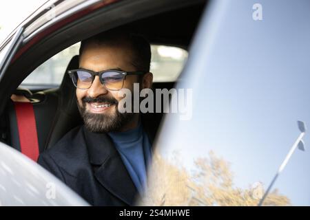 Cheerful Indian businessman looks away while smiling in his car, dressed in winter attire, reflecting a joyful commuting experience. Stock Photo