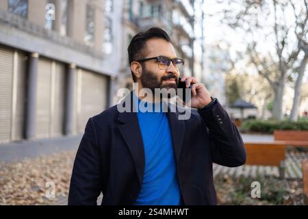 Indian businessman in winter clothing using a mobile phone in an urban park setting, looking away from the camera. Stock Photo