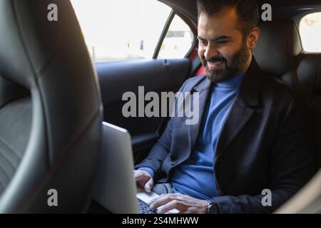 A happy Indian man dressed in professional attire works on his laptop while sitting in the backseat of a car, showcasing a modern, mobile business lif Stock Photo
