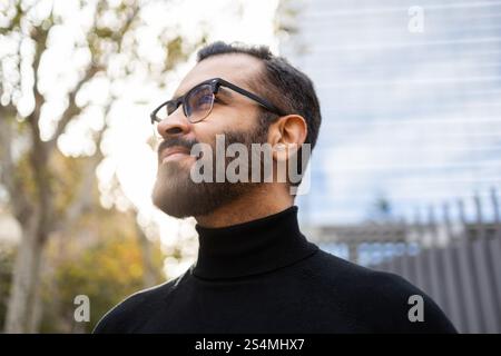 Portrait of a mature Indian businessman dressed in a black turtleneck, wearing glasses, and soaking up the sunny ambiance on a winter day, outdoors wi Stock Photo