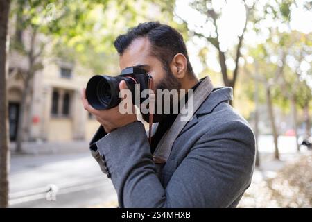An Indian man, dressed as a photographer in a fashionable winter coat, takes a photo with a professional camera on a leafy city street. Stock Photo