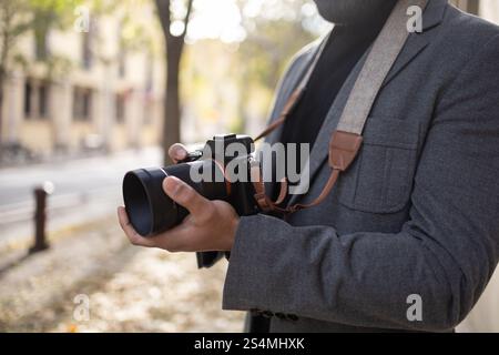 Cropped unrecognizable man, dressed as a photographer with a stylish gray jacket and a camera strap, holds a digital camera, focusing intently on capt Stock Photo
