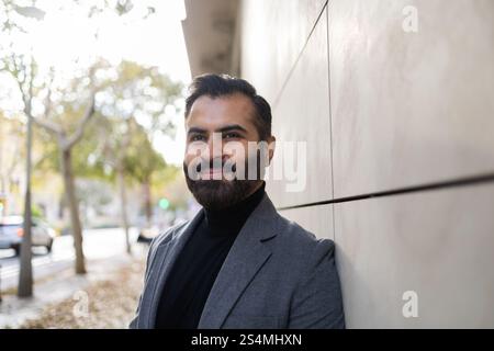 A cheerful Indian man in winter clothing, featuring a grey coat and black turtleneck, standing outdoors with a slight smile, looking away Stock Photo