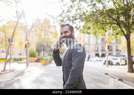 Cheerful Indian man in winter business attire, on a call using a mobile phone on a sunny urban street, embodying a modern, active lifestyle. Stock Photo