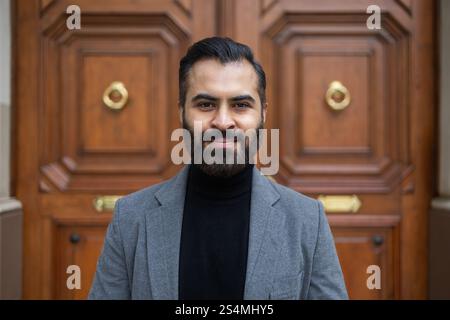 Portrait of a confident Indian businessman in a grey coat and black turtleneck, standing in front of a classic wooden door, looking directly at the ca Stock Photo