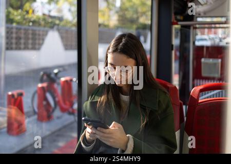 A young woman wearing glasses and a green coat looks at her smartphone while riding public transportation The dimly lit interior and window reflection Stock Photo
