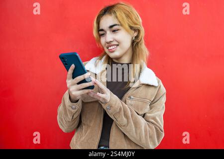 A mixed race woman smiles as she uses a smartphone, against a vibrant red background She is wearing a casual brown jacket with a white sheepskin colla Stock Photo