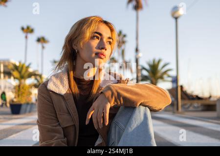 Mixed race woman sitting near the beach, gazing thoughtfully at the sunset She is surrounded by palm trees, creating a serene and contemplative atmosp Stock Photo