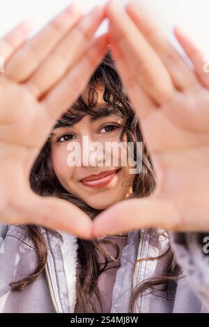 A young Latin woman with curly hair smiles through a hand frame, enjoying a sunny day Her casual style and natural charm embody a carefree and lively Stock Photo