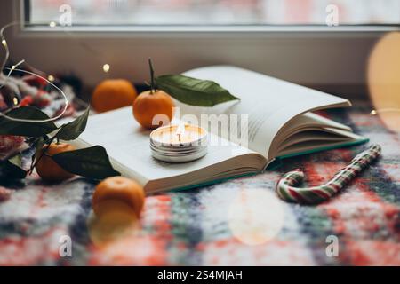 Christmas setting featuring a burning candle on an open book, surrounded by oranges and holiday lights, reflecting warmth and seasonal cheer. Stock Photo