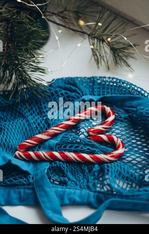 A blue string bag overflowing with red and white striped candy canes, placed alongside a pine branch adorned with twinkling lights, creates a cozy win Stock Photo