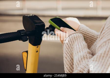 Cropped unrecognizable woman scans QR code with a smartphone to rent an electric scooter. The focus is on her hands holding both the phone and scooter Stock Photo