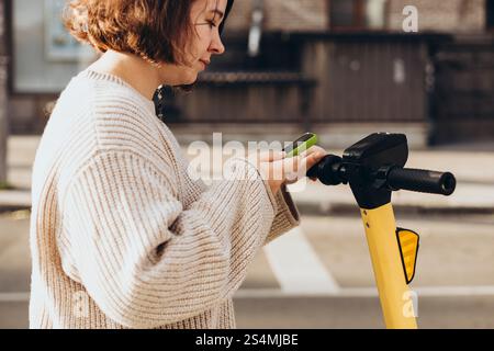 A woman is engaged in scanning a QR code with her smartphone to rent an electric scooter, standing outdoors on a sunny day. The scene captures her as Stock Photo