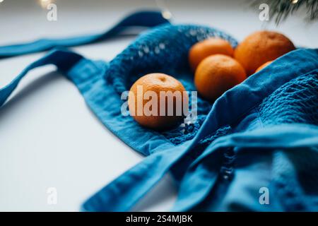 Close-up view of ripe tangerines neatly tucked inside a vibrant blue string bag, set against a soft, blurred background with subtle lighting. Stock Photo