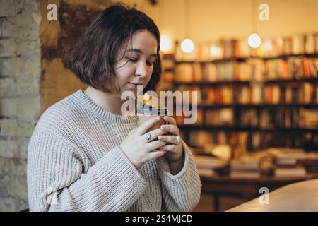 A woman in a soft sweater holds a takeaway coffee cup while enjoying a peaceful moment in a cozy bookstore Bookshelves in the background create a warm Stock Photo