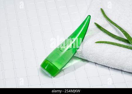 A green aloe vera gel tube rests next to aloe leaves on a white fluffy towel, all arranged on square bathroom tiles. Stock Photo