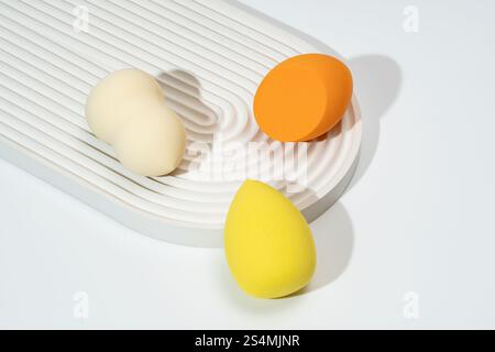 A trio of colorful cosmetic sponges in orange, yellow, and cream are arranged on a textured, white surface The minimalist composition highlights the b Stock Photo