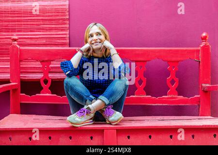 A woman wearing a blue plaid shirt sits cross-legged on red wooden stairs, smiling gently and looking away against a vibrant pink wall. Stock Photo