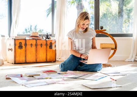 A woman sits on a bright home floor, absorbed in her laptop, with papers scattered around her. Sunlight streams through large windows. Stock Photo