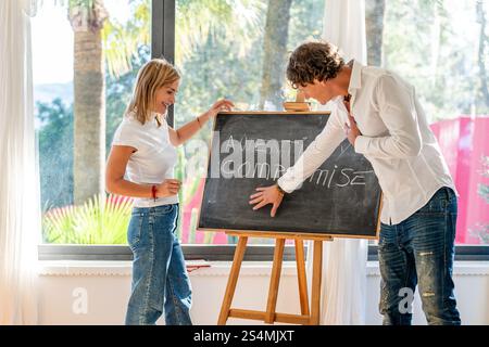 A young man and woman collaborate cheerfully around a chalkboard, standing in a bright room with a decorative background. Stock Photo