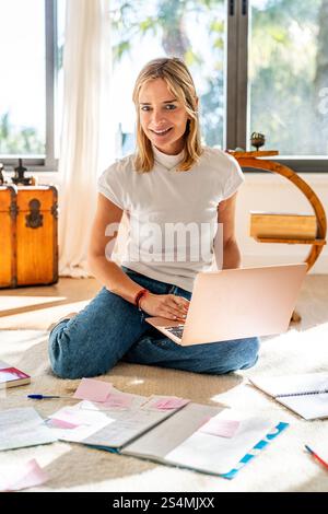 Cheerful woman working from home, sitting cross-legged on the floor with a laptop and paperwork. She is looking directly at the camera and smiling, il Stock Photo