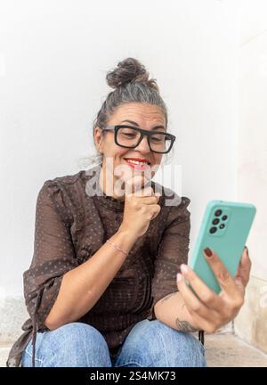 A woman with glasses uses a smartphone for a video call, focusing on learning sign language She is seated against a light background, exuding enthusia Stock Photo