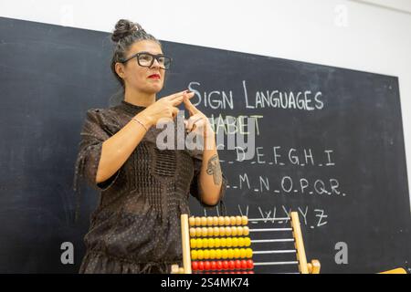 An instructor teaching sign language in a classroom, standing in front of a chalkboard with the alphabet written on it An abacus is visible in the for Stock Photo