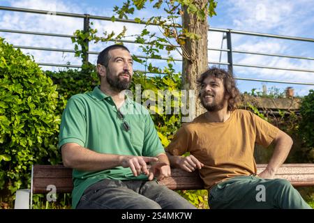 Two men sit on a bench surrounded by lush greenery One man, who is blind, appears relaxed and engaged in conversation with his companion, highlighting Stock Photo