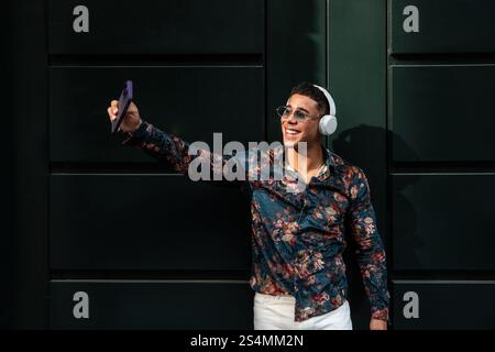 A young latin man in a floral shirt, wearing sunglasses and headphones, smiles while taking a selfie against a dark green wall. The sun highlights his Stock Photo