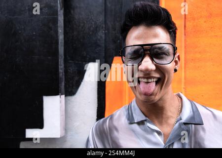 A young latin man wearing oversized sunglasses sticks out his tongue in a playful manner while standing in front of a vibrant orange and black wall, e Stock Photo