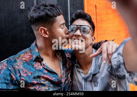 Latin gay couple taking selfie while embracing warmly. One partner kisses the other's cheek, while both are smiling under a bright orange background. Stock Photo