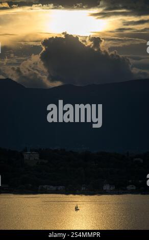 A sailboat glides on shimmering Lake Geneva under a dramatic sunset sky Dark silhouettes of distant hills frame the scene, with clouds adding depth an Stock Photo