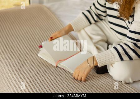 A person in a cozy, striped sweater sits cross-legged on a bed with a textured blanket, holding an open blank notebook, creating a peaceful and relaxe Stock Photo